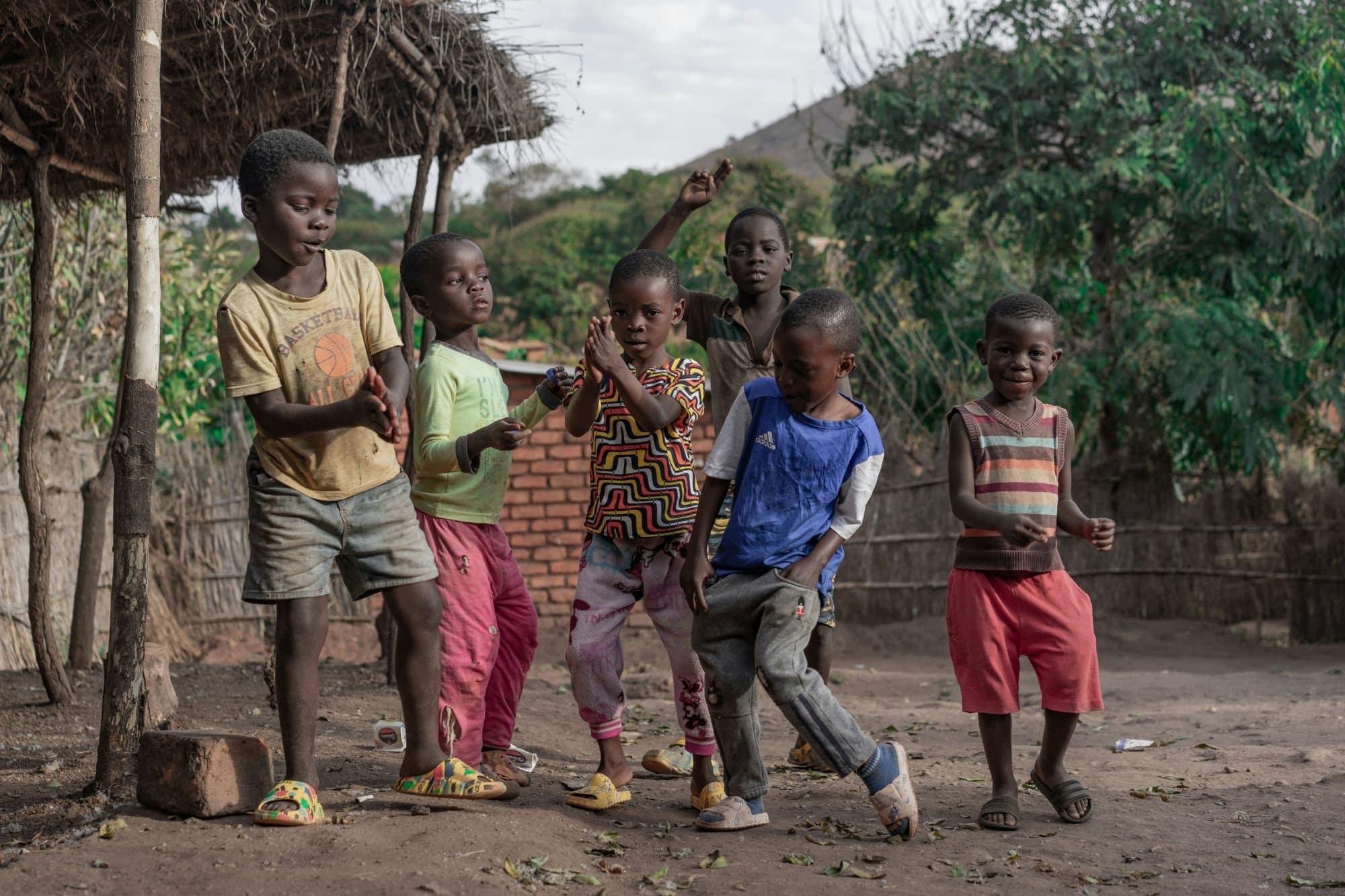 Children playing together in a Kupanda Collective community, near a traditional thatched-roof structure